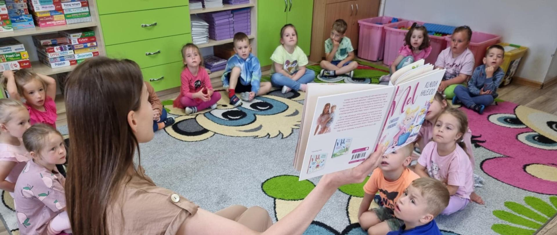 
Grupa przedszkolaków siedzi w kole na przedszkolnym dywanie. Mama jednego z dzieci czyta książeczkę.