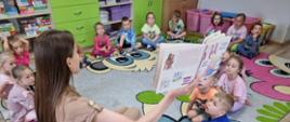 Grupa przedszkolaków siedzi w kole na przedszkolnym dywanie. Mama jednego z dzieci czyta książeczkę.