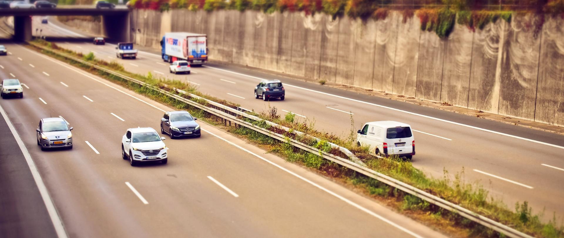Trzy pasmowa droga szybkiego ruchu. Po drodze jadą samochody. W tle zdjęcia widoczny most.