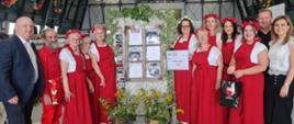 Zdjęcie przedstawia fotografię członków Koła Gospodyń Wiejskich „Polne Kwiaty” z Rutek, uwiecznioną na tle udekorowanego stoiska z dyplomami i polnymi kwiatami. Kobiety oraz jeden z mężczyzn mają na sobie charakterystyczne, wyraziste czerwone stroje (fartuchy i koszule) oraz wianki na głowach. Po bokach grupy stoją oficjalni goście w eleganckich ubraniach.