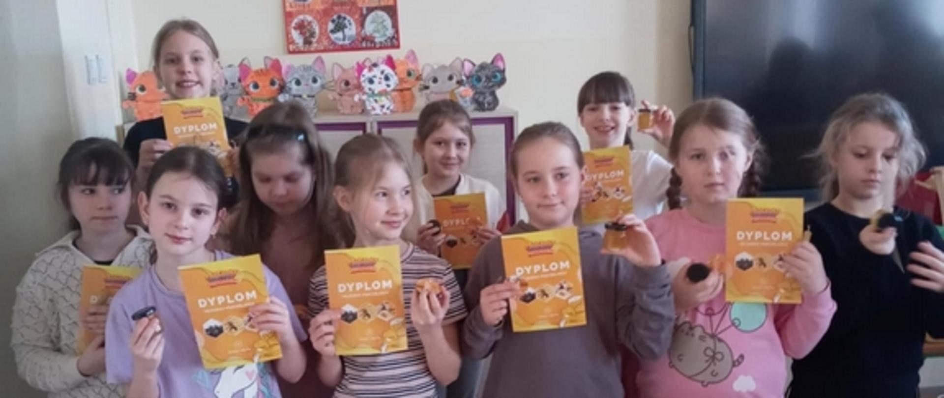 Grupka dziewczynek z klasy drugiej stoi w sali lekcyjnej trzymając w jednej ręce dyplomy uczestnictwa w warsztatach pszczelarskich, a w drugiej mały słoiczek z miodem.