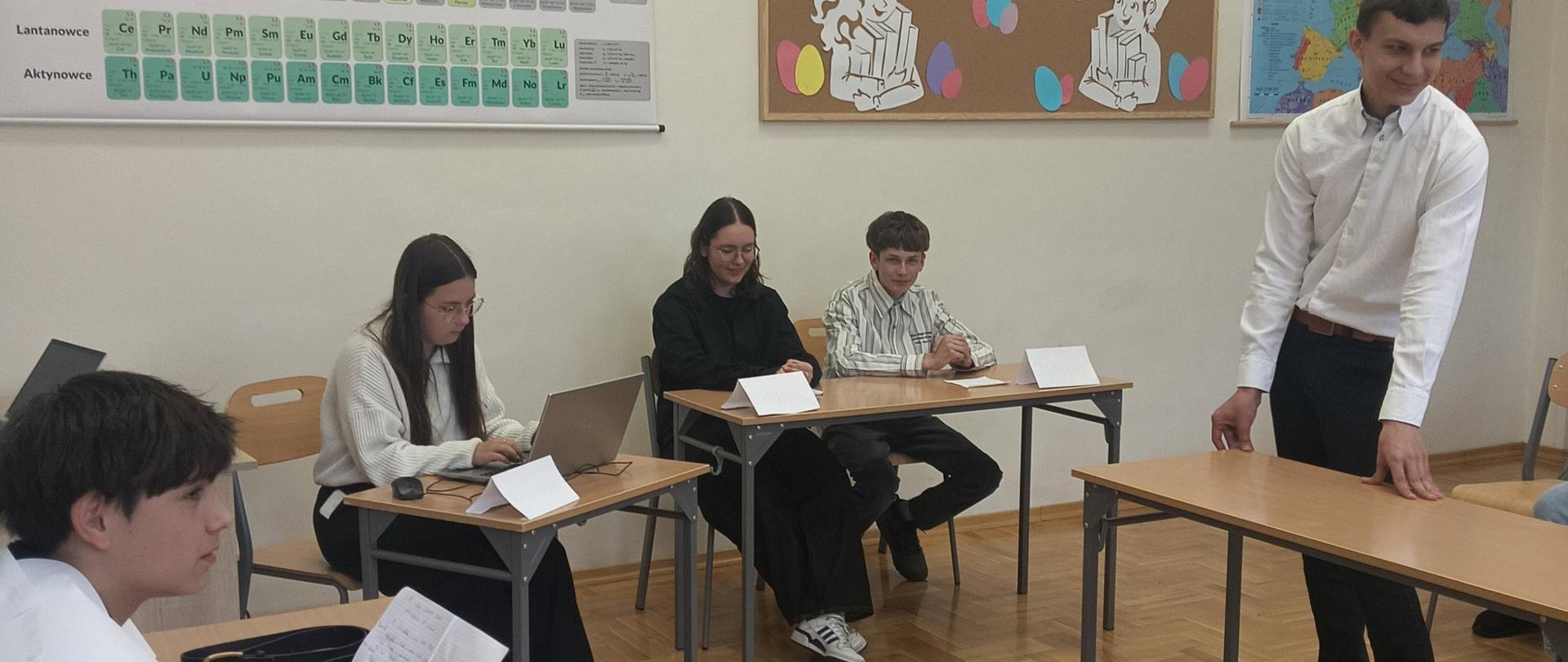 Na zdjęciu widać salę lekcyjną podczas szkolnej debaty lub prezentacji. Kilka osób siedzi przy ustawionych w podkowę ławkach, a jedna osoba stoi po prawej stronie, prawdopodobnie zabierając głos lub przygotowując się do wypowiedzi. Uczniowie mają przed sobą kartki, notatki i laptopy, co sugeruje przygotowanie do dyskusji lub konkursu.
Na ścianach wiszą pomoce naukowe: duża tablica z układem okresowym pierwiastków, kolorowa gazetka z napisem „WIOSNA” oraz mapa Europy. Wnętrze jest jasne i schludne, z drewnianą podłogą i prostym szkolnym wyposażeniem. Atmosfera wygląda na spokojną, skupioną i oficjalną, ale jednocześnie przyjazną.