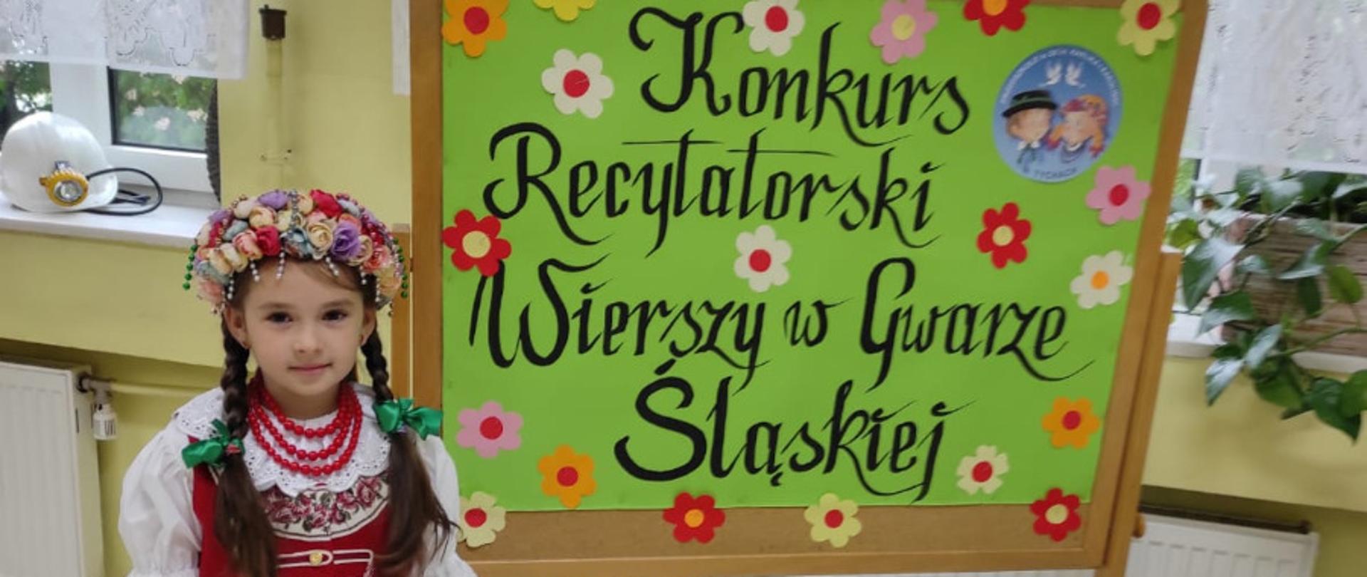 Dziewczynka w stroju śląskim przy zielonej tablicy z napisem konkurs recytatorski wierszy w gwarze śląskiej ozdobionej kwiatkami