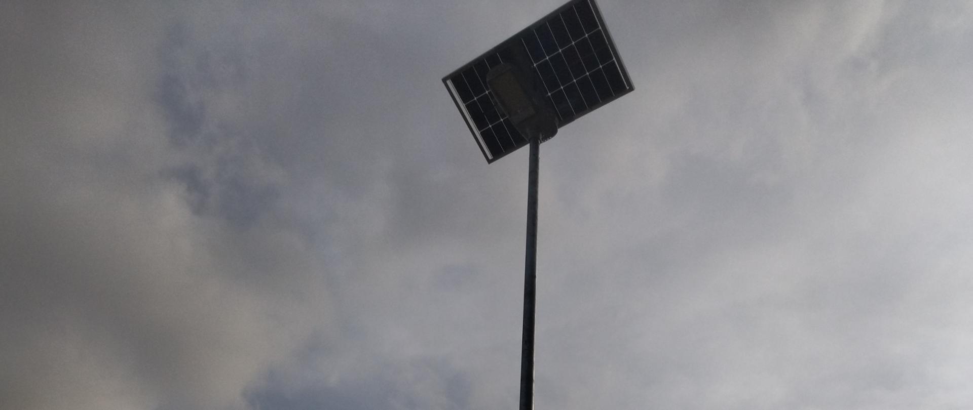 Obraz przedstawia wysoki metalowy słup, na którego szczycie zamontowany jest panel słoneczny. Słup znajduje się na trawiastym terenie z drzewami w tle. Do słupa przymocowana jest tablica informacyjna, a po prawej stronie znajduje się kolejna tablica. Niebo jest zachmurzone, a przez obraz przechodzi pozioma linia energetyczna.