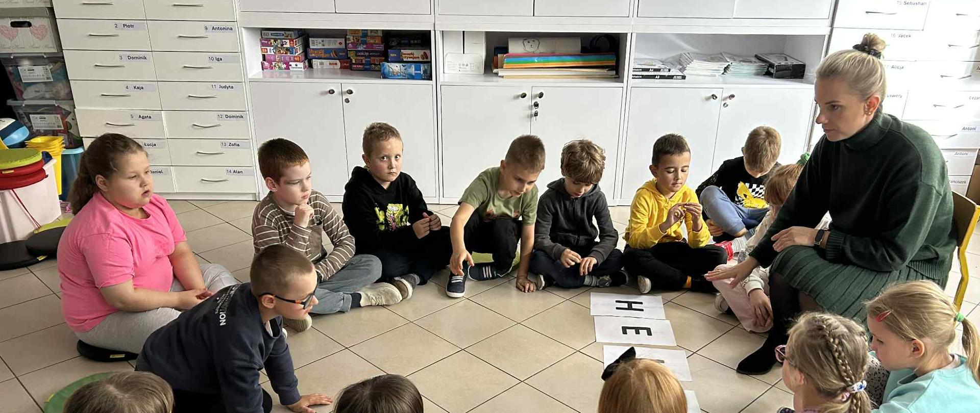 Dzieci siedzą w okręgu na podłodze. Między nimi siedzi kobieta na krześle. Po środku leżą kartki z dużymi literami