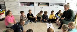 Dzieci siedzą w okręgu na podłodze. Między nimi siedzi kobieta na krześle. Po środku leżą kartki z dużymi literami