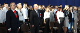 W sali stoi grupa ludzi, a wśród nich mężczyzna w eleganckim garniturze.