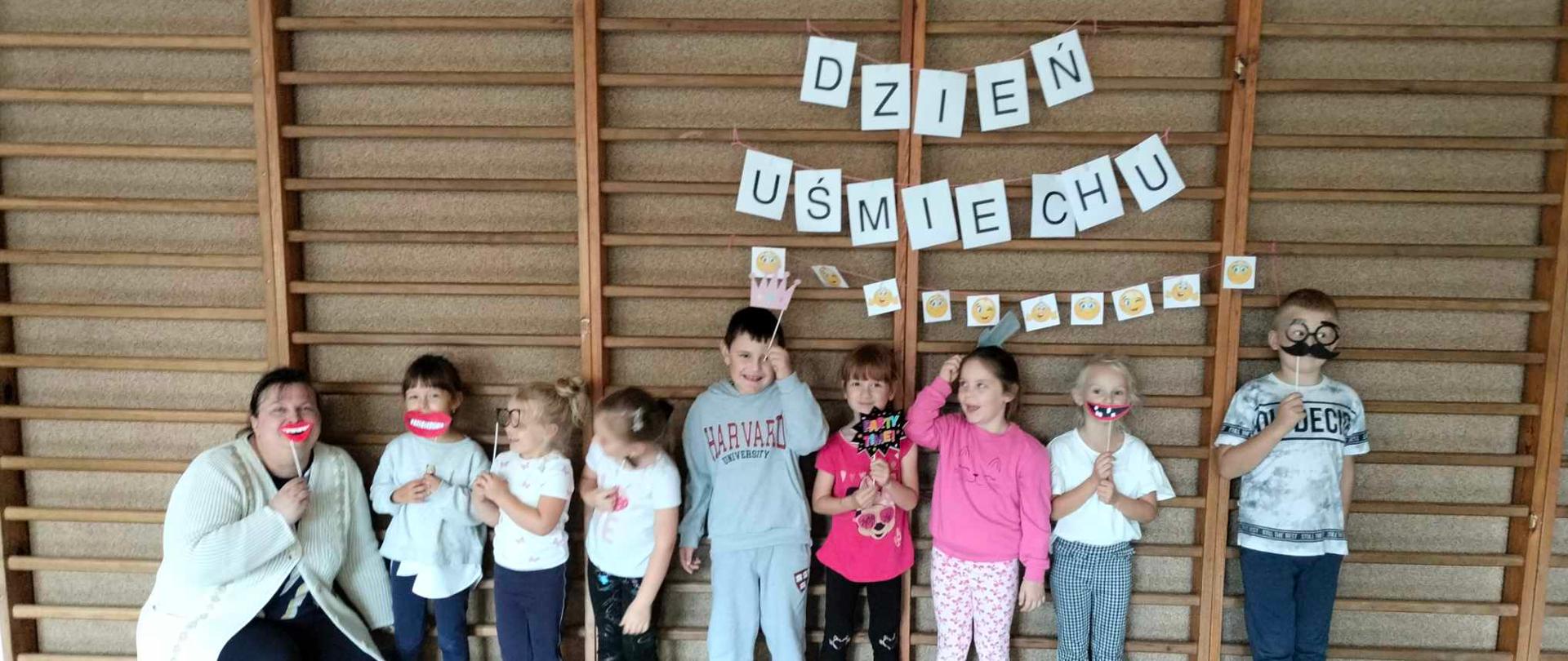Ośmioro przedszkolaków oraz Pani pozują do zdjęcia z papierowymi gadżetami. Za nimi na drabinach widnieje napis DZIEŃ UŚMIECHU oraz obrazki przedstawiające uśmiechnięte buźki.