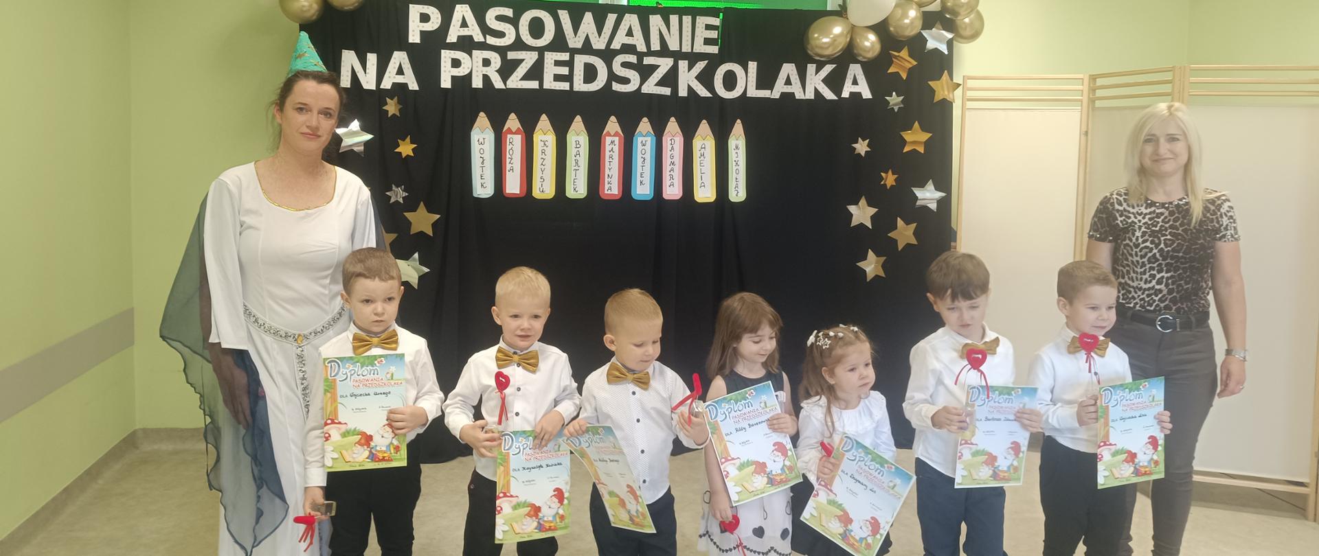 Zdjęcie przedstawia grupę dzieci w wieku przedszkolnym stojących obok siebie w rzędzie. Chłopcy mają ciemne spodnie i białe koszule, dziewczynki białe bluzeczki i ciemne spódniczki. Po lewej stronie stoi pani w stroju wróżki, po prawej pani w ciemnych spodniach i bluzeczce w panterkę. Za nimi scenografia - ciemne tło na którym przypięto napis Pasowanie na przedszkolaka, kredki z imionami dzieci, kolorowe gwiazdki i baloniki. Ściany sali są w kolorze zielonym. 