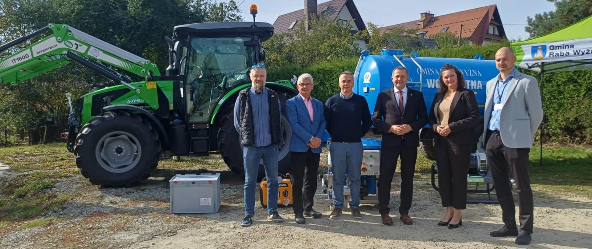 Grupa osób pozuje do wspólnego zdjęcia na tle ciągnika, beczkowozu z wodą pitną i sprzętu technicznego.