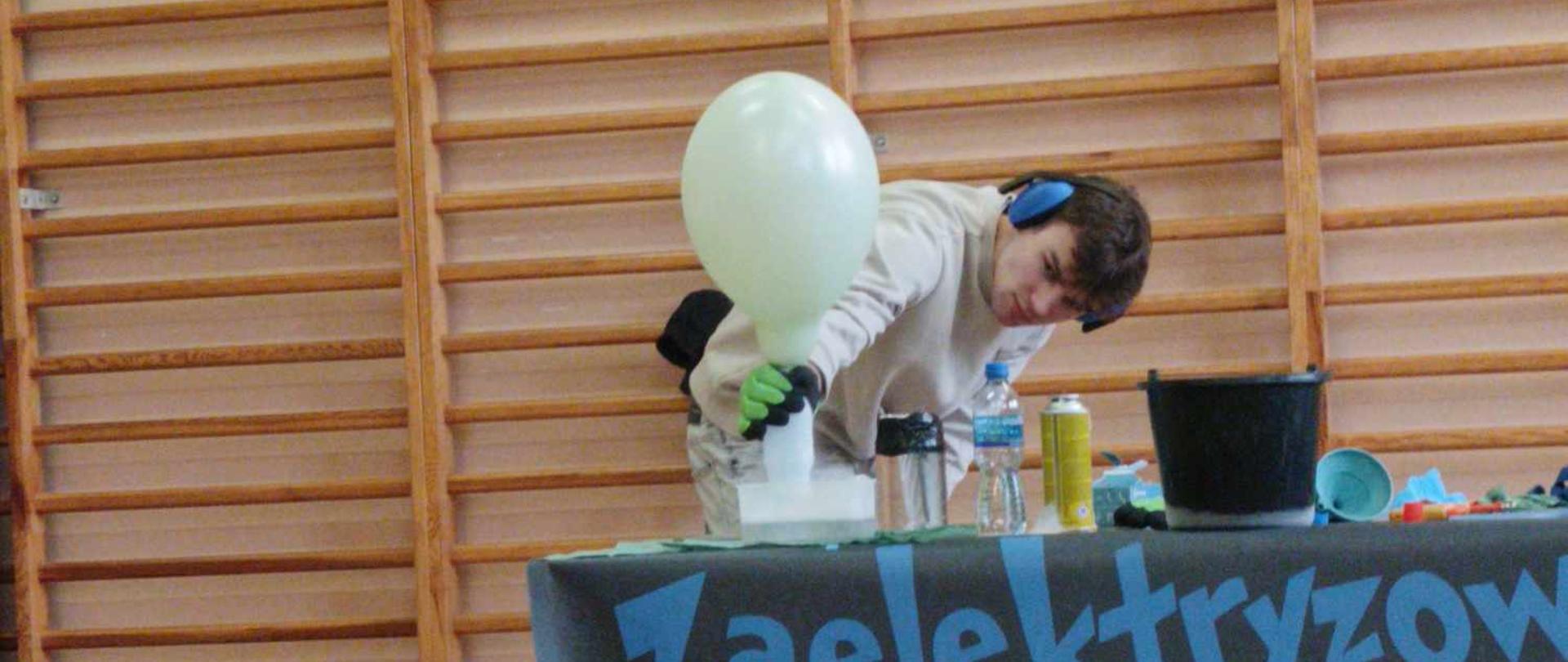 Mężczyzna w niebieskich słuchawkach ochronnych trzyma napompowany balon nad butelką z białym gazem