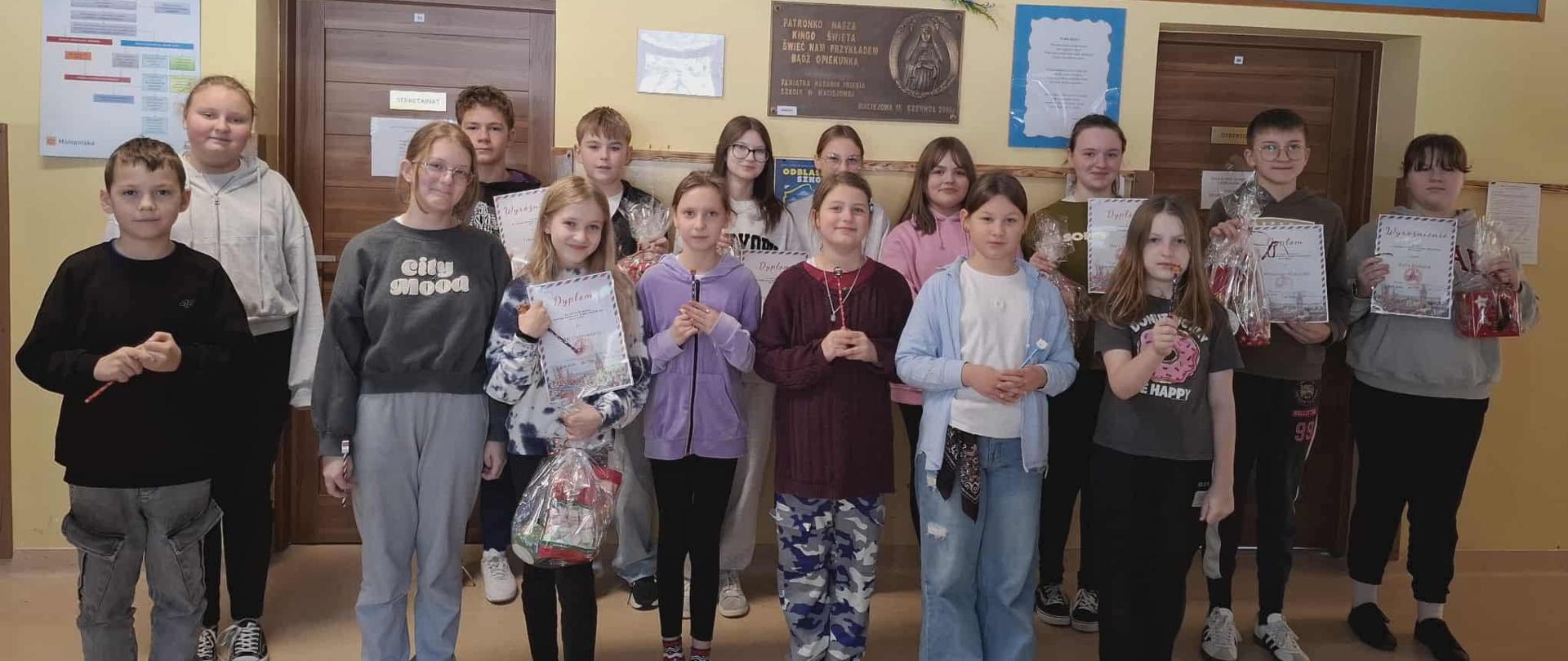 Uczniowie biorący udział w konkursie stoją pod ścianą na której wisi tablica pamiątkowa z nadania imienia szkole, kwiaty i obraz patronki szkoły świętej Kingi, zwycięzcy trzymają w ręku nagrody oraz dyplomy
