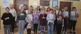 Uczniowie biorący udział w konkursie stoją pod ścianą na której wisi tablica pamiątkowa z nadania imienia szkole, kwiaty i obraz patronki szkoły świętej Kingi, zwycięzcy trzymają w ręku nagrody oraz dyplomy
