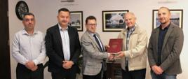 Reprezentanci stron podpisujących umowę pozują do wspólnego zdjęcia podczas przekazania podpisanych dokumentów. Starosta przekazuje teczkę z dokumentami i ściska dłoń osoby reprezentującej spółkę wykonawcy. W tle gabinet i wiszące na ścianach obrazki z miejscami na terenie powiatu sokołowskiego.