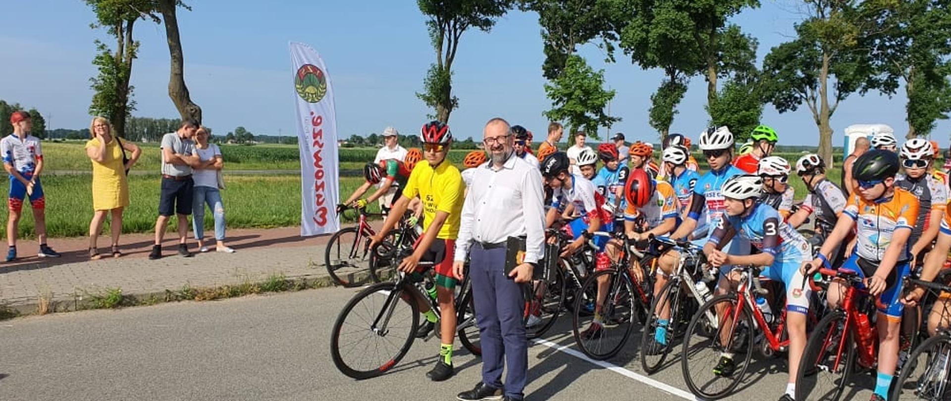 Zdjęcie przedstawia Starostę Przasnyskiego w białej koszuli i granatowych spodniach stojącego obok siedzącego na rowerze kolarza w żółtej koszuli i czerwonym kasku. Za nimi na linii startowej stoi reszta uczestników wyścigu.