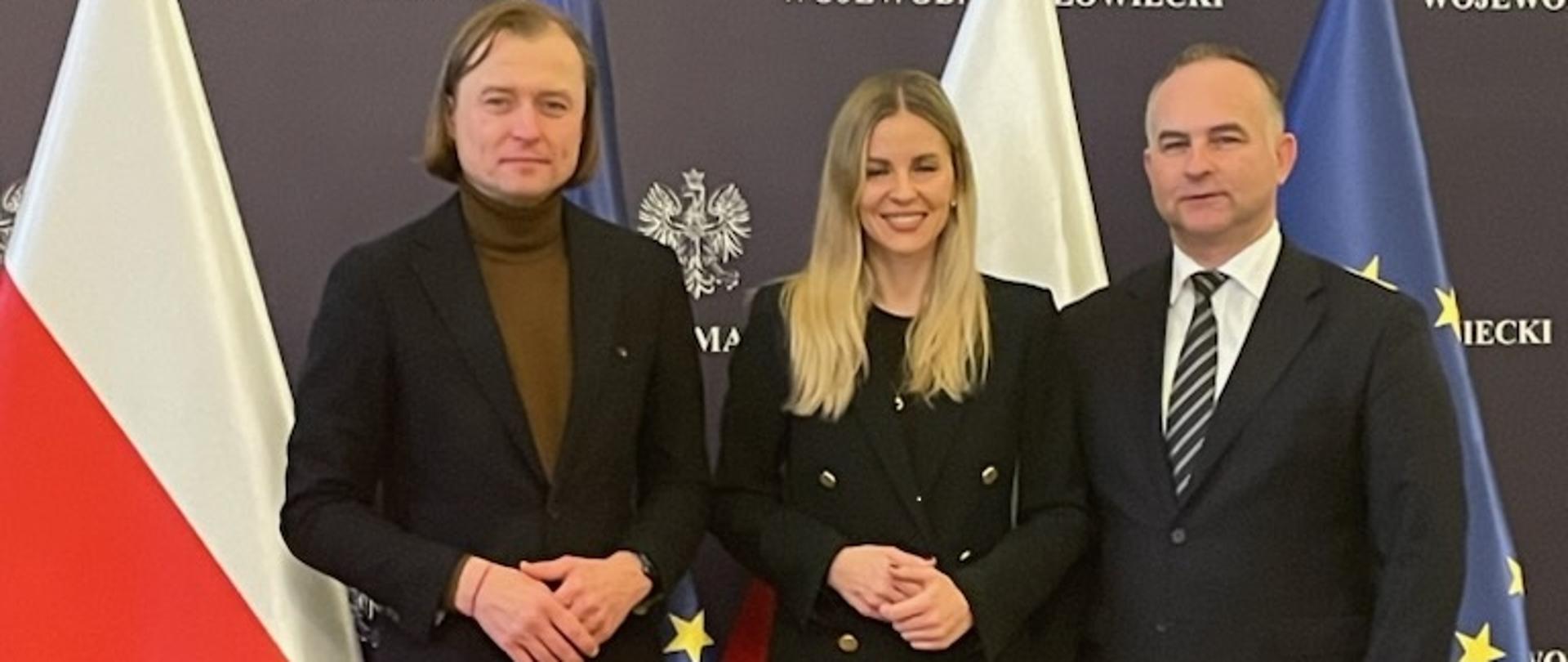 trzy osoby stojące- dwóch mężczyzn i kobieta-, za nimi flagi: od lewej: na przemian Polski i Unii Europejskiej
