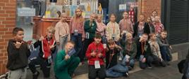 Grupa dzieci sto i kuca przed szklanym wejściem do budynku. Dzieci mają zawieszone na czerwonych smyczkach identyfikatory
