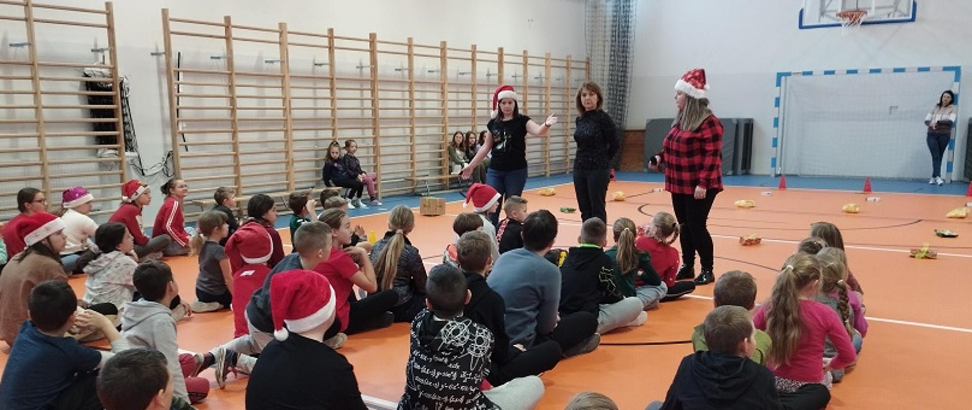 Uczniowie siedzą po turecku na sali gimnastycznej słuchając instrukcji nauczyciela.