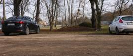 Zdjęcie przedstawia betonowe boisko, na którym stoją dwa samochody. Dalej poza boiskiem rosną drzewa