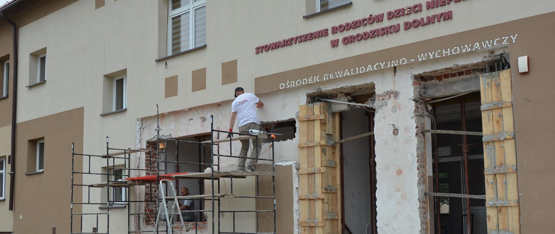 bezowy budynek w remoncie brak drzwi wejściowych i okna, na rusztowaniu robotnik z narzędziami , 