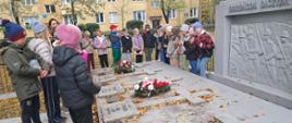 Grupa dzieci stoi obok poziomego pomnika z czterema krzyżami. Na nimi leżą dwie wiązanki z biało czerwonymi kwiatami
