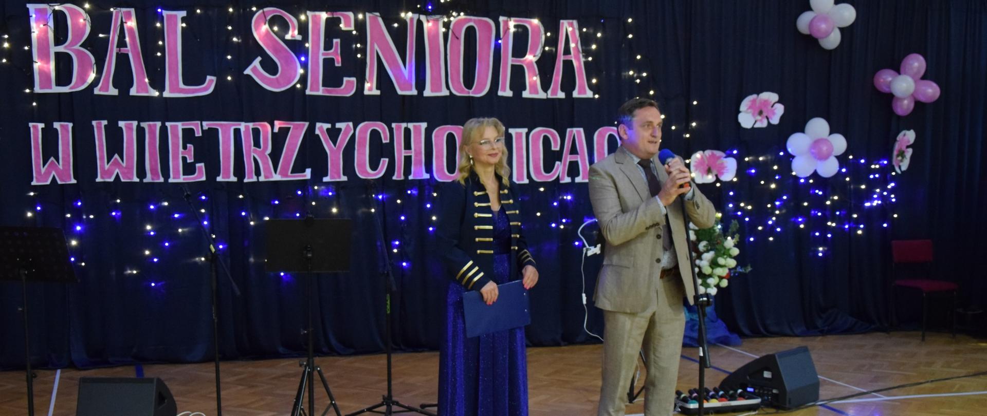 Kobieta i mężczyzna stoją na scenie z mikrofonami podczas oficjalnego otwarcia Balu Seniora w Wietrzychowicach. W tle widoczny jest duży napis wydarzenia oraz dekoracje świetlne.