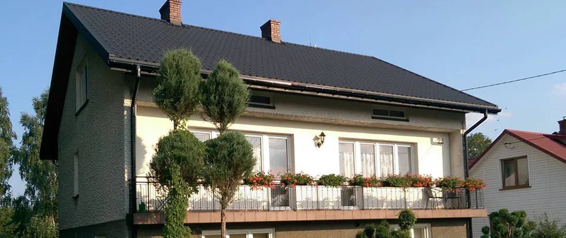 Piętrowy dom z długim balkonem przez szerokość całego domu, przed domem ogród.