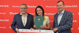 Trzy osoby stoją na tle czerwonej ścianki z powtarzającym się napisem „Mazowsze”. Od lewej: wójt gminy Pokrzywnica Adam Dariusz Rachuba, skarbnik gminy Magdalena Lusa oraz wicemarszałek województwa mazowieckiego Wiesław Raboszuk. Trzymają duży, symboliczny czek.