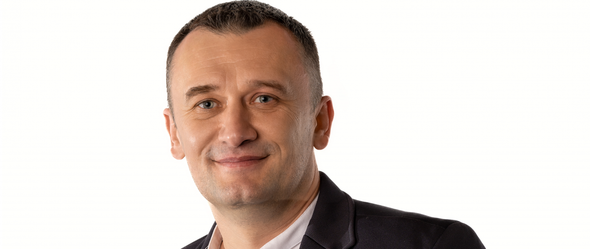Szatyn o niebieskich oczach i śniadej karnacji, uśmiechnięta twarz. Ubrany w granatową marynarkę i białą koszulę ze stójką, która ma rozpięty jeden guzik od góry