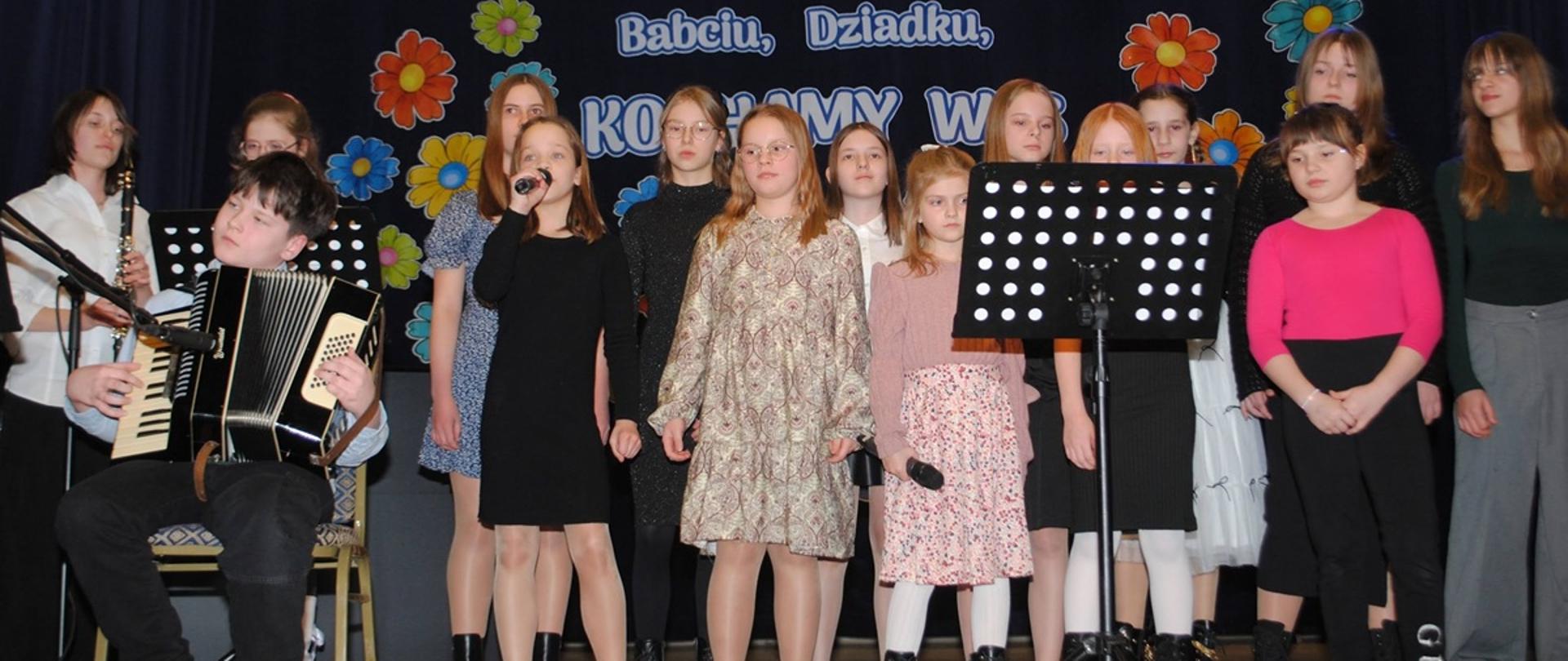 dziewczynki stoją na scenie i śpiewają jeden chłopak siedzi i gra na akordeonie 