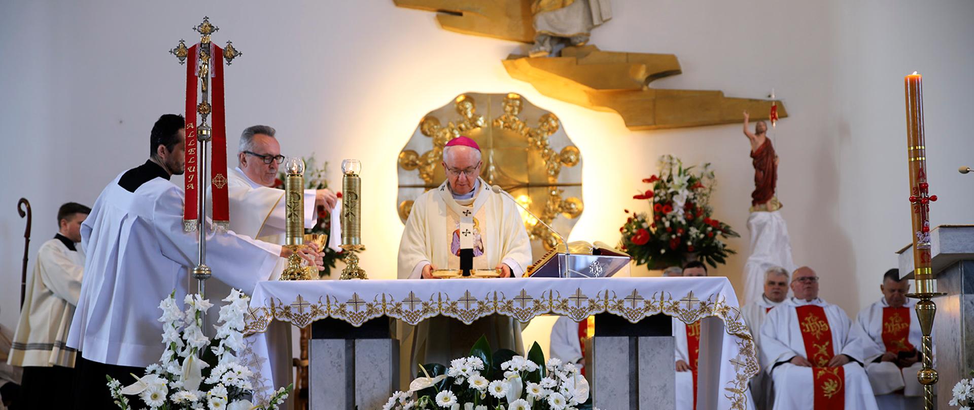 Zdjęcie przedstawia ozdobiony kwiatami stół ofiarny przy którym Arcybiskup prowadzi liturgię. W tle znajduje się podświetlony ołtarz.