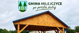 Zdjęcie przestawia drewniana wiatę w tle budynek świetlicy w Rogaczach
