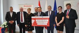 Grupa osób stoi razem, trzymając duży symboliczny czek z napisem „Dofinansowanie z RPO WM 2014–2020” na kwotę 9 838 602,07 zł. W tle widoczne są flagi Polski i Unii Europejskiej oraz dwa herby umieszczone na ścianie.