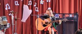 Na scenie znajduje się dziewczyna. Uczennica siedzi na krześle i gra na gitarze. Ubrana jest w czarną marynarkę i czerwoną spódnicę. Za nią, na czerwonym tle widnieje napis: ,,VIII POWIATOWY KONKURS PIOSENKI OBCOJĘZYCZNEJ AUGUSTÓW 2025".