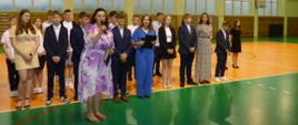 Grupa kilkunastu uczniów wraz z Panią Dyrektor stoją w jednej linii na zielonej płycie boiska, w tle okna hali sportowej. Pani Dyrektor przemawia do mikrofonu.