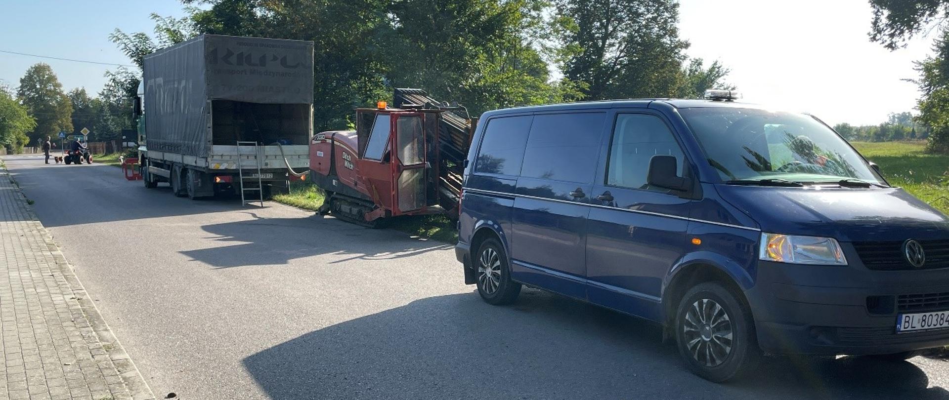Ulica i chodnik, na poboczu stoją dwa samochody oraz maszyna do wodociągów.