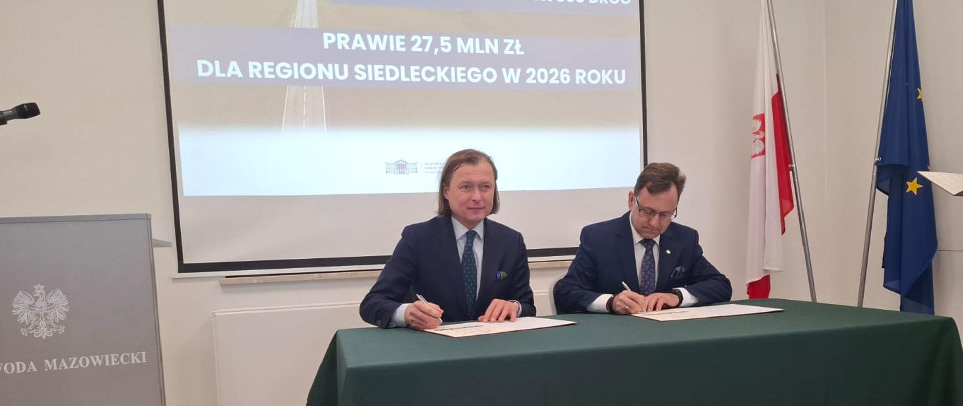 Starosta i Wojewoda siedzą przy stole nakrytym obrusem i podpisują umowę.