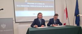 Starosta i Wojewoda siedzą przy stole nakrytym obrusem i podpisują umowę.
