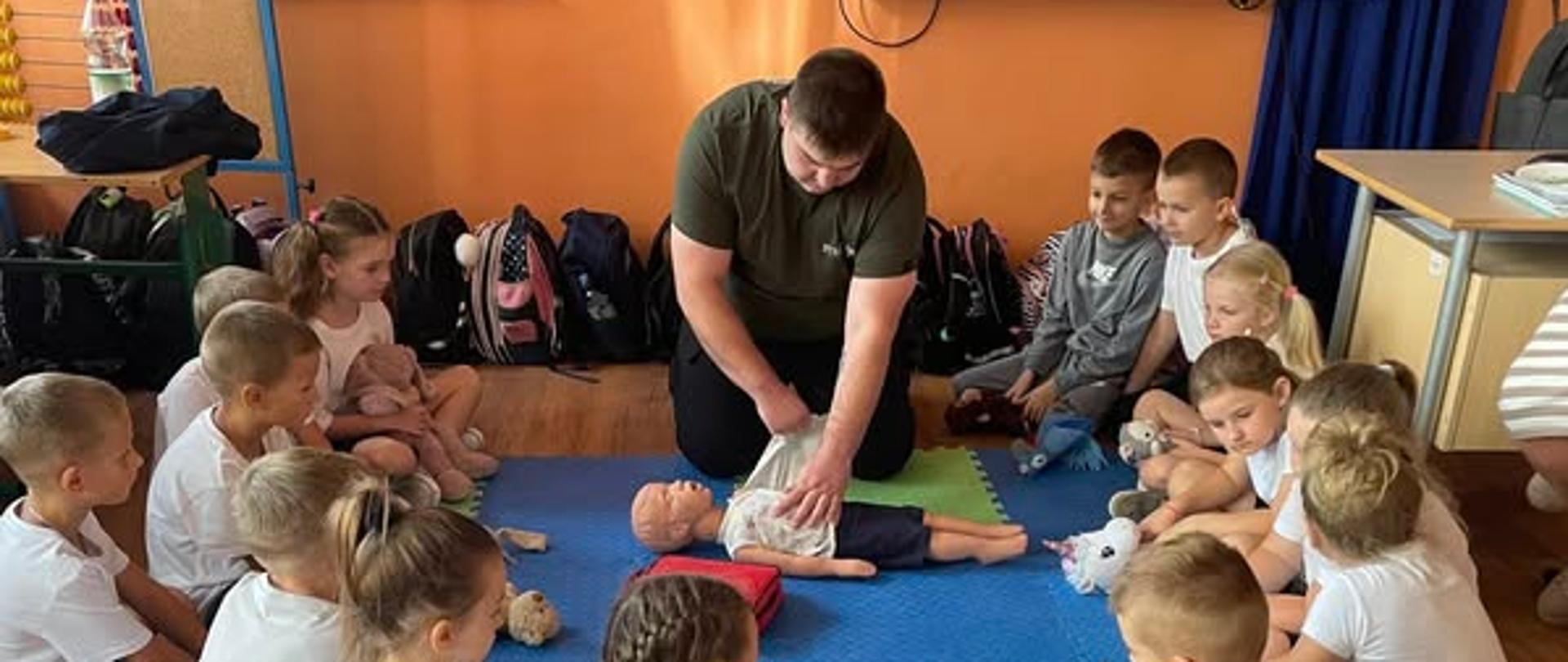 Ratownik wykonuje masaż serca na manekinie, dzieci siedzące w kręgu przyglądają się tej czynności