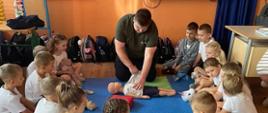 Ratownik wykonuje masaż serca na manekinie, dzieci siedzące w kręgu przyglądają się tej czynności