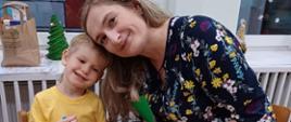 Chłopczyk i jego mama siedzą przy stole, wspólnie tworząc świąteczne ozdoby - papierową torbę z bożonarodzeniowym akcentem