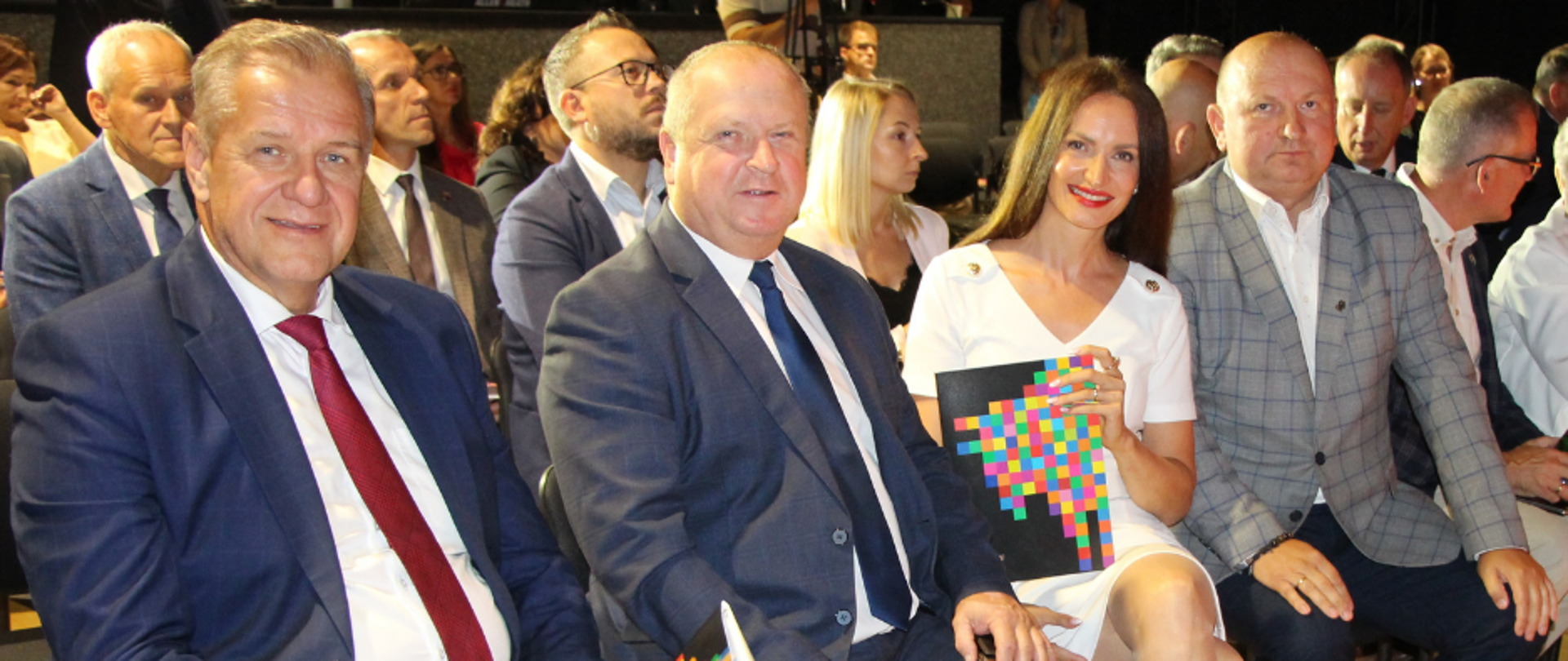 Przedstawiciele samorządu siedzą w pierwszym rzędzie podczas wydarzenia. Trzej mężczyźni w garniturach oraz kobieta w białej sukience trzymają pamiątkowe materiały z kolorową grafiką.