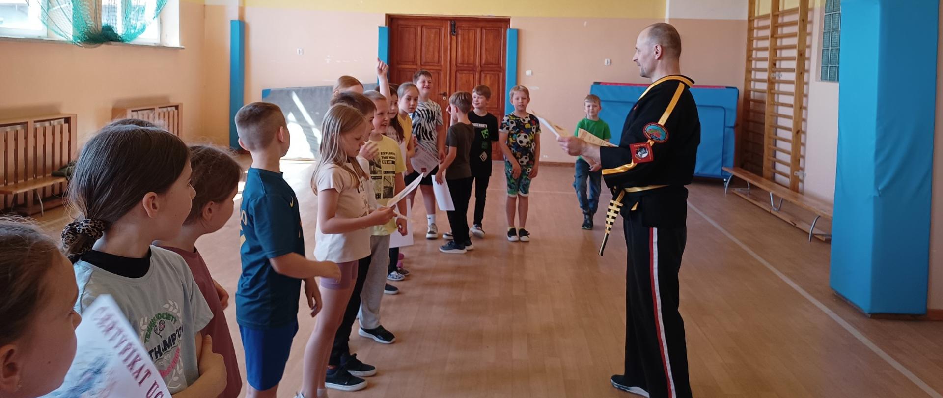 Dzieci ustawione w rzędzie na sali gimnastycznej. Na przeciwko dzieci stoi mężczyzna ubrany w czarne kimono