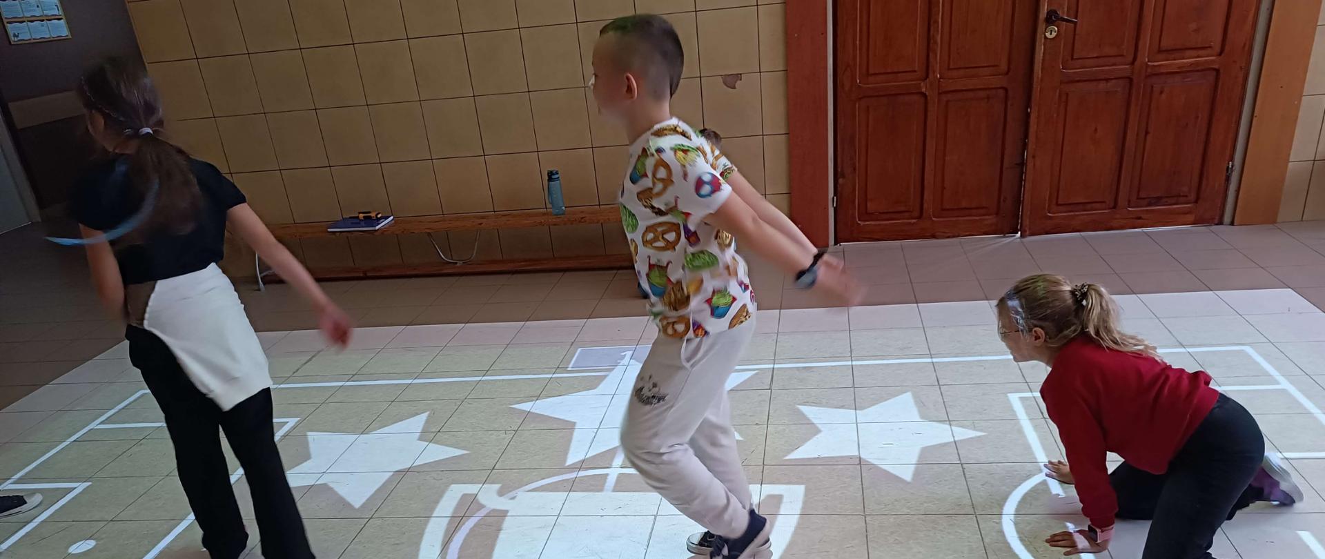 Troje dzieci. Jedna z dziewczynek na kolanach. Dwoje innych chodzi. Na podłodze wyświetlany biały obraz boiska, pucharu i gwiazd