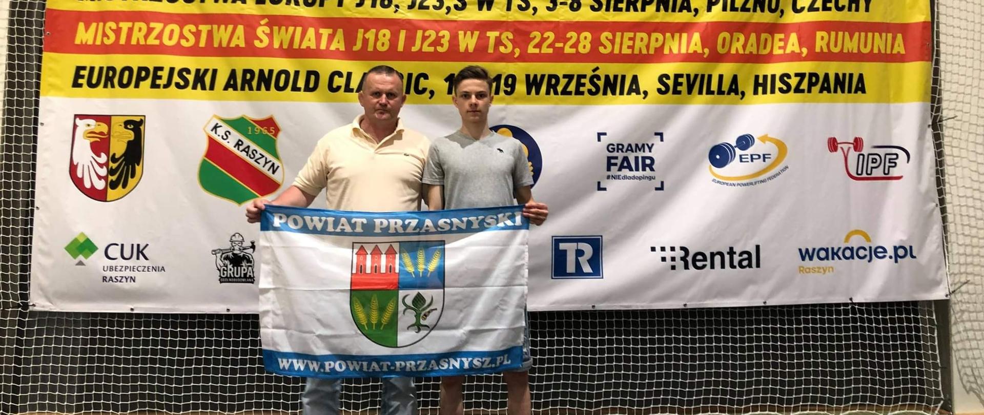 Zdjęcie przedstawia trenera Cezarego Pykałę oraz zawodnika Jakuba Bońkowskiego wspólnie trzymających flagę z herbem Powiatu Przasnyskiego. W tle widnieje biała siatka, do której przymocowano baner promujący wydarzenie: Puchar Polski w trójboju siłowym oraz eliminacje do Mistrzostw Europy, Mistrzostw Świata oraz Europejskiego Arnold Classic.