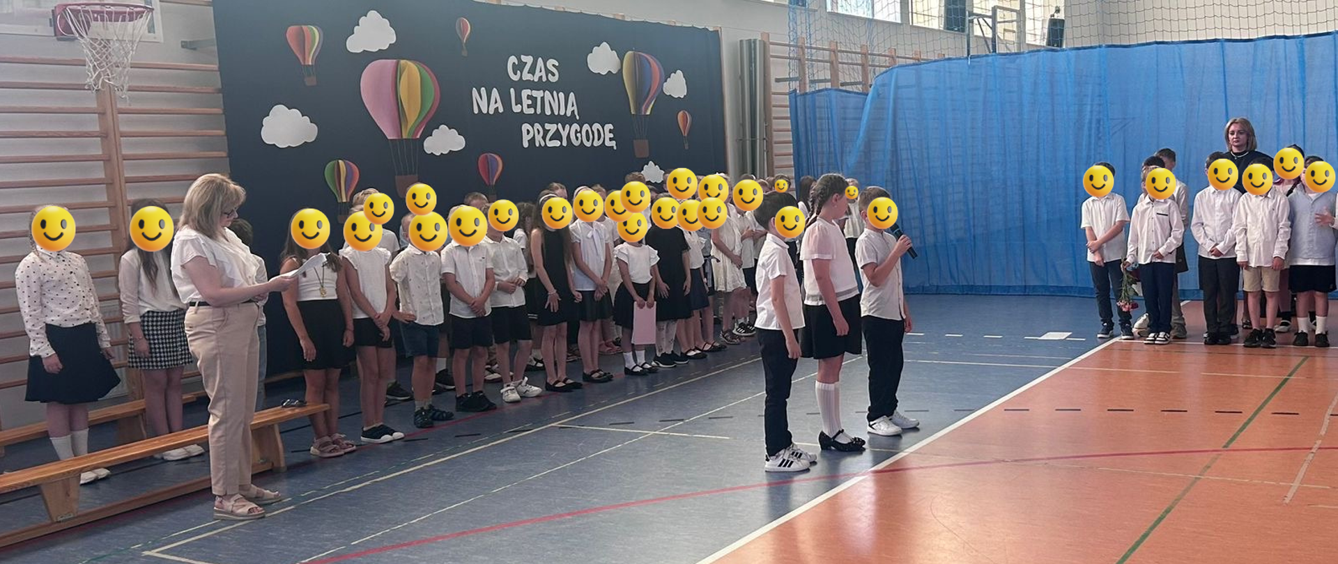 Sala gimnastyczna, po lewej stronie widać ławki. Przy nich w dwóch rzędach stoją uczniowie. Obok nich stoi pani. Przed nimi w równej linii stoi trójka dzieci, jedno z nich trzyma mikrofon. W tle widać niebieską zasłonę, do której przyczepiony jest biały napis, chmury i balony. Po prawej stronie widać dzieci ustawione w rzędzie. Za nimi stoi pani. W tle widać niebieską zasłonę.