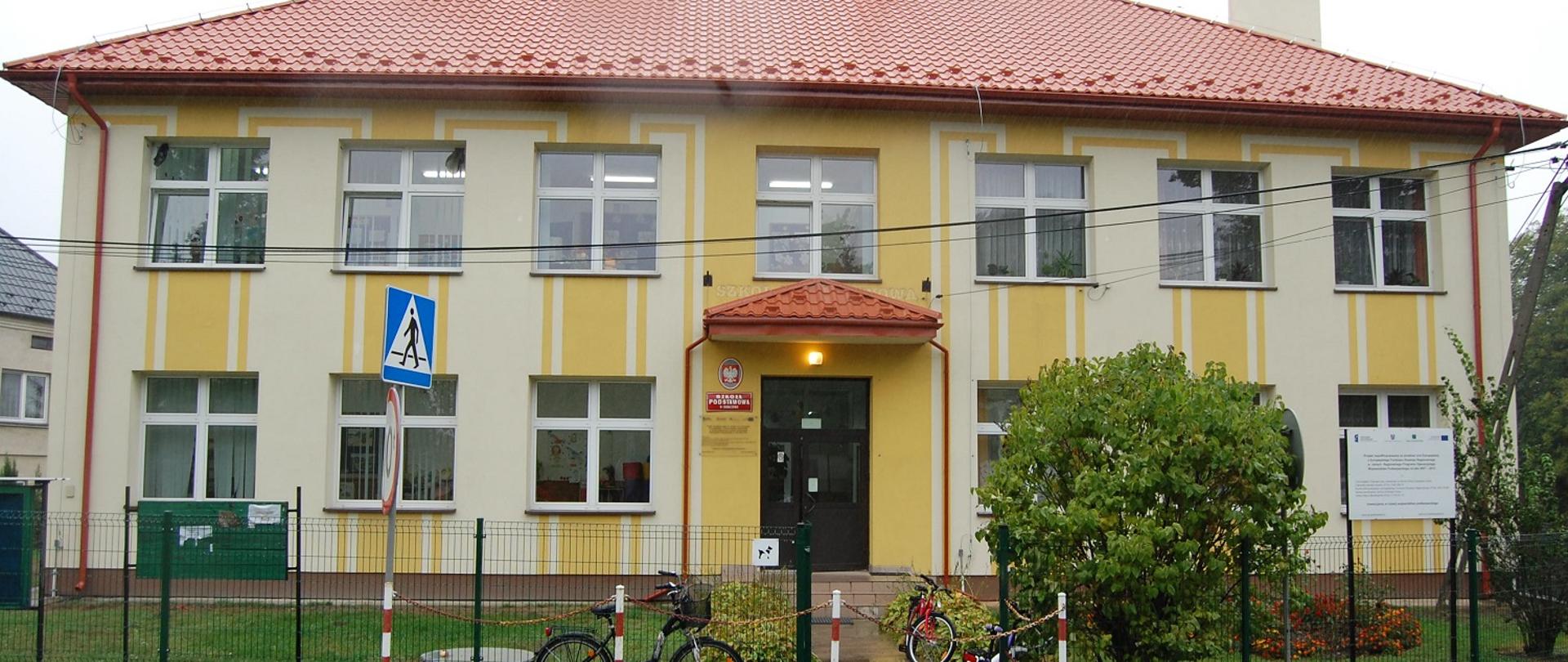 Biało - żółty budynek szkolny z czerwonym dachem. Przed budynkiem trzy rowery oparte o zielone ogrodzenie. 