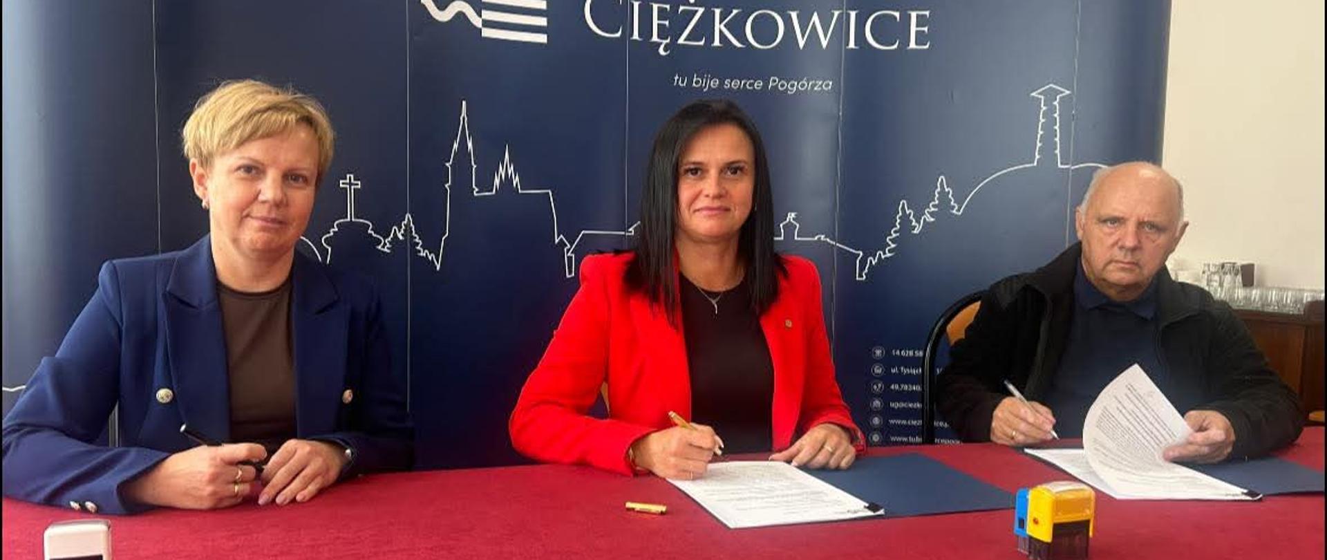 Trzy osoby siedzące na tle granatowej ściany z napisem gmina Ciężkowice