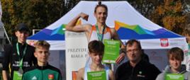 Olek Dziemiańczyk trzyma dyplom, stoi na podium pozuje do zdjęcia z trenerem i pozostałymi laureatami biegu