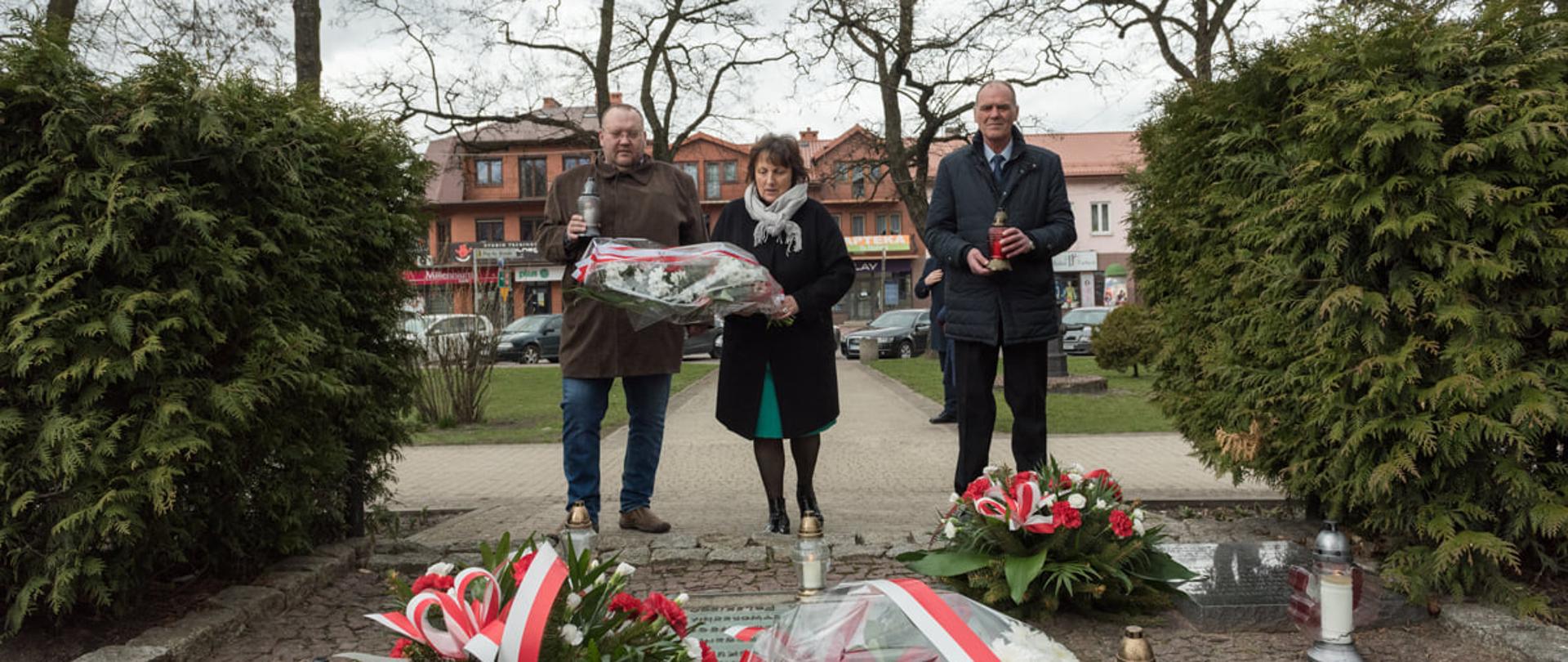 Widok z perspektywy pomnika. Kwiaty składa starosta sokołowski Elżbieta Sadowska. Po jej prawej stronie stoi przewodniczący Rady Powiatu Jacek Odziemczyk, który trzyma w ręku biały znicz, po lewej wicestarosta Ryszard Domański z czerwonym zniczem. Zbliżają się do pomnika, u podstawie którego widać inne biało - czerwone wiązanki.